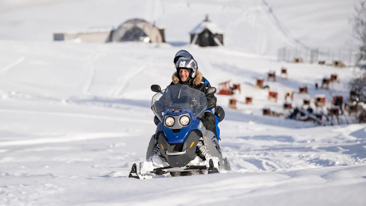 To personer på en blå snøscooter kjører på snødekt terreng, med Arctic Domes og hundegårder i bakgrunnen.