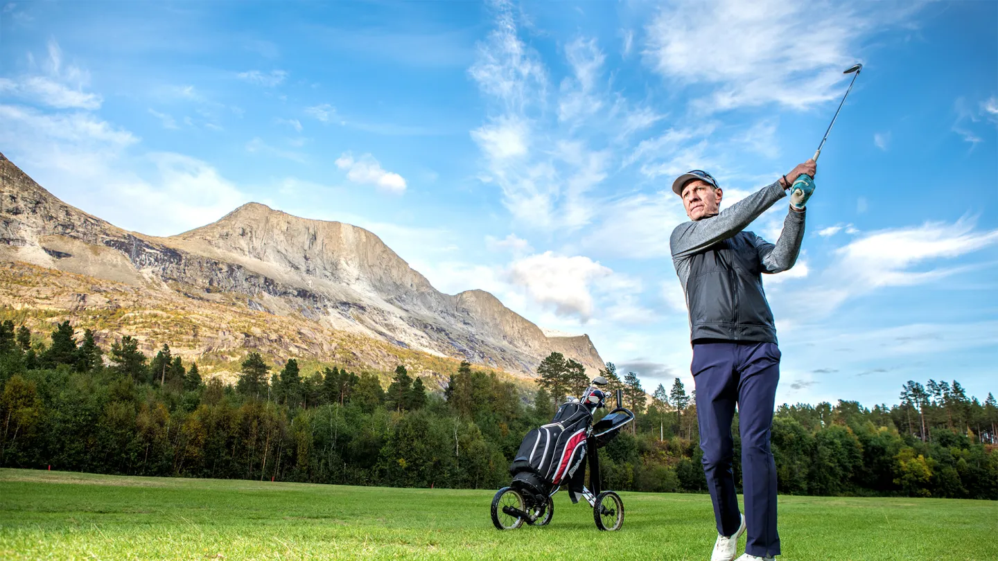 Golfspiller slår et slag på grønn gressbanen i Skjomen med golftralle og bag, fjell og skog under blå himmel.