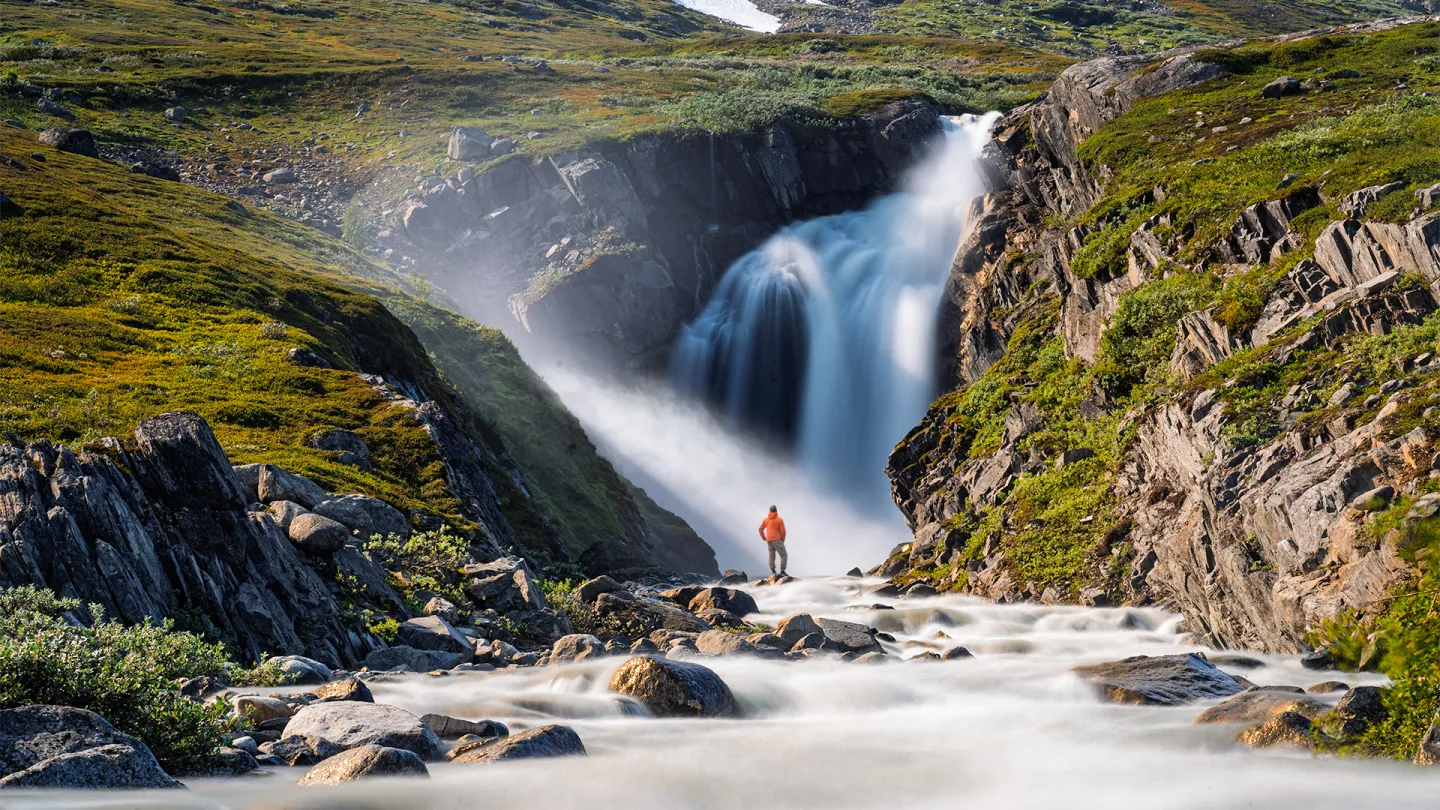 En person i oransje jakke står ved en elv foran en stor foss i et grønt, steinete fjellandskap.
