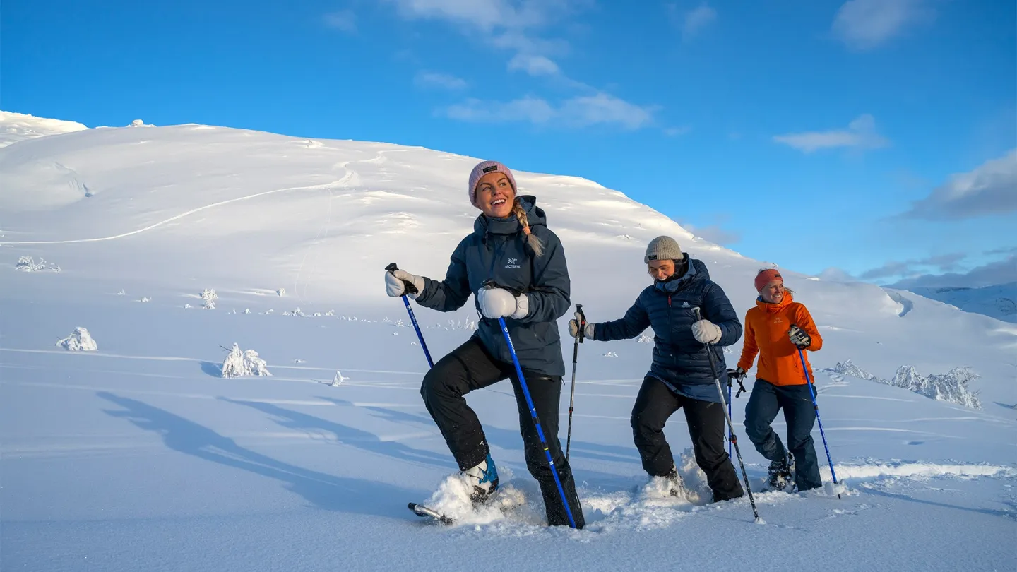 Tre personer går med truger og staver i dyp snø i Narvikfjellet under blå himmel med spredte skyer.