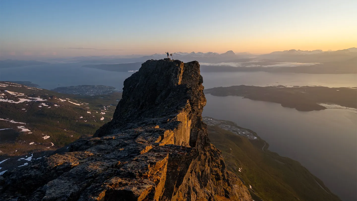 En person står med armene løftet på Rombakstøtta over fjord og øyer i varmt kveldslys.