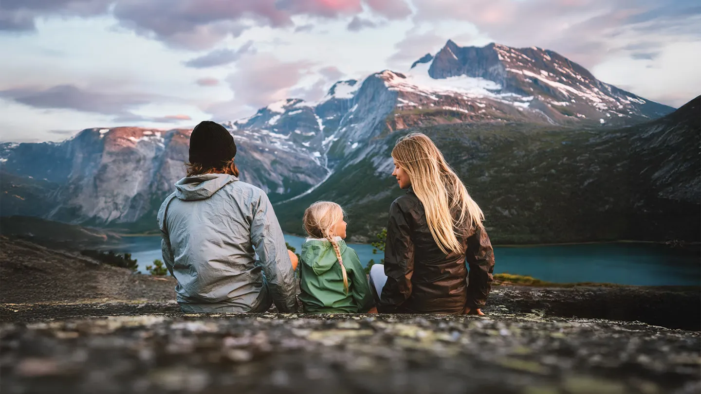 Tre personer sitter på en steinrygg og ser mot et fjellmassiv med snøflekker og en innsjø under rosa skyer.