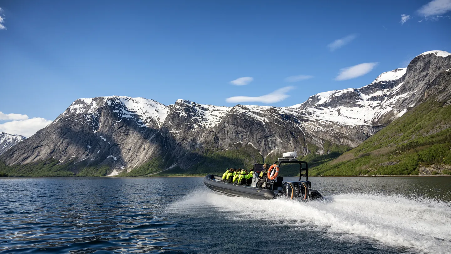 En ribbåt med passasjerer i redningsvester suser over fjorden med snødekte fjell og blå himmel i bakgrunnen.