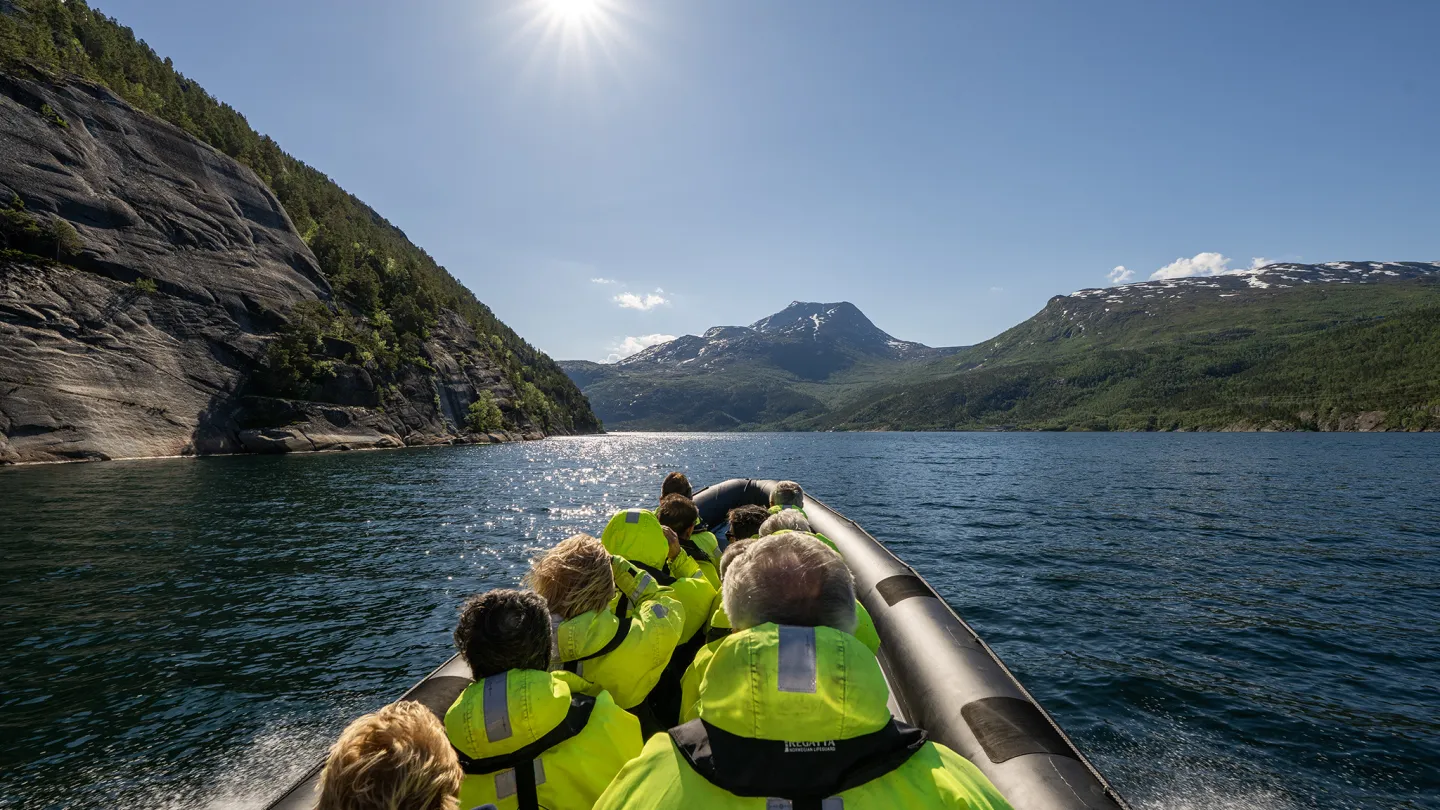Passasjerer i gule flytedrakter sitter i en RIB-båt på blå fjord, med fjell og sol på himmelen.