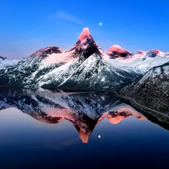 Snødekt Stetind i rosa vinterlys speiler seg i en stille fjord, med månen over og en vei langs bredden.