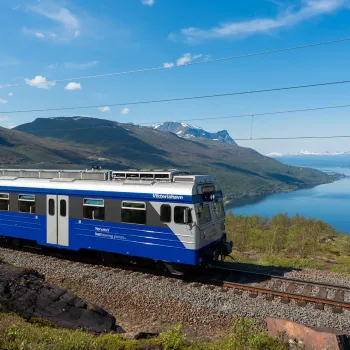 Arctic Train langs Ofotbanen med fjord, grønne fjell og blå himmel i bakgrunnen.