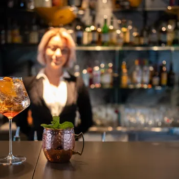To drinker på en bardisk foran en bartender, med flasker og glass i hyller bak.