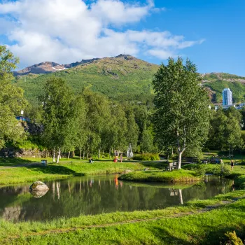Den frodige parken i Elvedalen i Narvik med dam og trær, sti og bro, med fjell og noen høye bygninger under blå himmel med skyer.