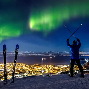 En skiløper står på en snødekt høyde med staver i kryss, mens nordlyset lyser over fjord og bylys.