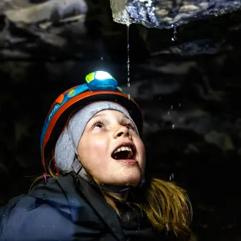 Person med hjelm og hodelykt står i en mørk grotte mens vann drypper fra steintaket over.