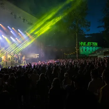 Stor folkemengde foran en utendørsscene om kvelden, med band på scenen og grønne lyskastere over publikum.