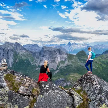 To kvinner på steinete fjellrygg ser utover grønne daler og spisse fjell under blå himmel med skyer.