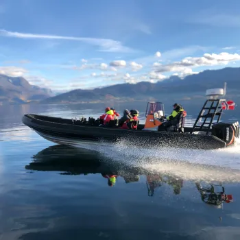 Svart RIB-båt med flere personer i redningsvester kjører raskt på speilblank Ofotfjord med fjell i bakgrunnen.