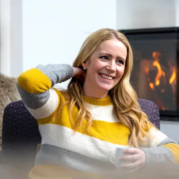 En kvinne i stripete genser sitter i en lenestol med kopp i hånden, foran en peis med flammer i bakgrunnen.