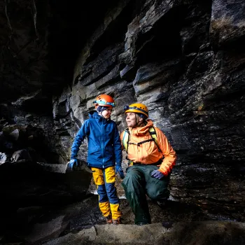 To personer med hjelmer og friluftsklær står på steiner i en mørk grotte med bratte fjellvegger.