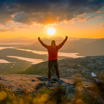 En person i rød jakke står på Verdenssvadet i Efjord med armene hevet, med innsjøer og fjell i midnattsol.