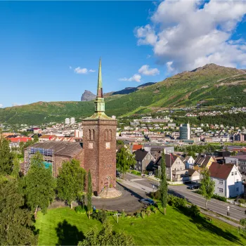 Narvik kirke med høyt spir i forgrunnen, omgitt av bolighus, grønt landskap og fjell under blå himmel.