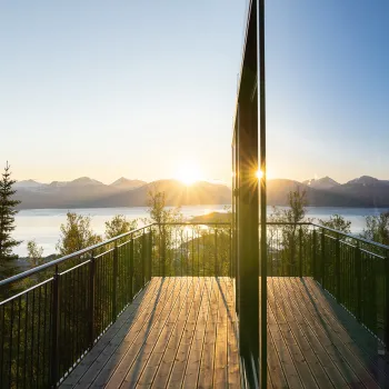 Utsikt fra treterrassen på Camp 291 i Narvik med rekkverk mot fjord og fjell, med lav sol og speiling i stort glassfelt.