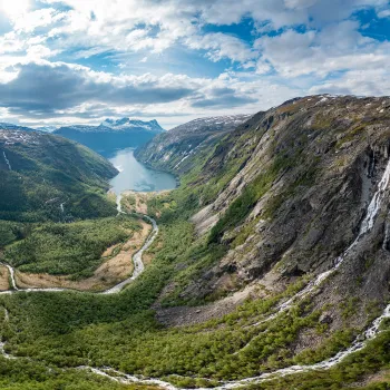 Panoramautsikt over Rombaksbotn med elv som slynger seg mot en innsjø, bratte fjell og foss under skyet himmel.