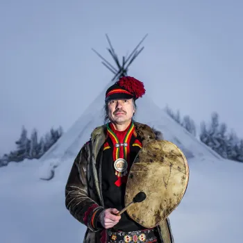 Sjaman Ronald Kvernmo i tradisjonell drakt holder en rund tromme og foran en snødekt lavvo i vinterlandskap.