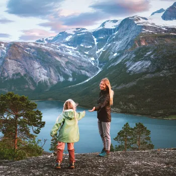 En voksen og et barn står på svaberget på Reinnesfjellet og ser utover en Skjlmenfjorden med skog og snødekte fjell i bakgrunnen.