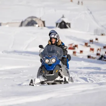 To personer på en blå snøscooter kjører på snødekt terreng, med Arctic Domes og hundegårder i bakgrunnen.