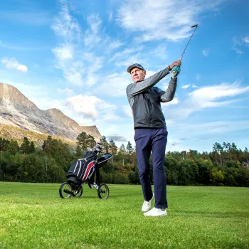 Golfspiller slår et slag på grønn gressbanen i Skjomen med golftralle og bag, fjell og skog under blå himmel.