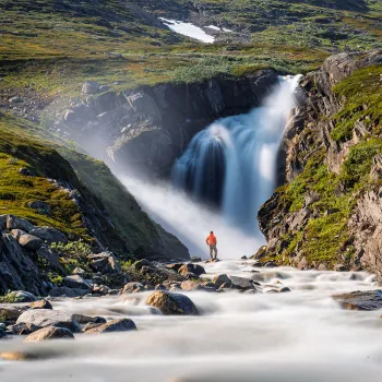 En person i oransje jakke står ved en elv foran en stor foss i et grønt, steinete fjellandskap.