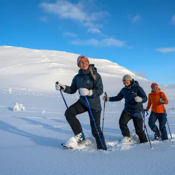 Tre personer går med truger og staver i dyp snø i Narvikfjellet under blå himmel med spredte skyer.