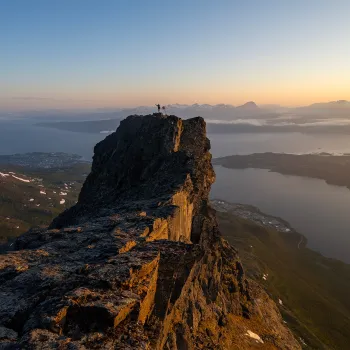 En person står med armene løftet på Rombakstøtta over fjord og øyer i varmt kveldslys.