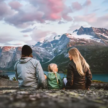 Tre personer sitter på en steinrygg og ser mot et fjellmassiv med snøflekker og en innsjø under rosa skyer.