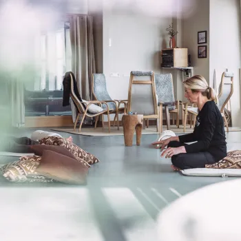 Person sitter på gulvmatte med puter i et lyst rom, med stoler i bakgrunnen og et tent lys på gulvet.