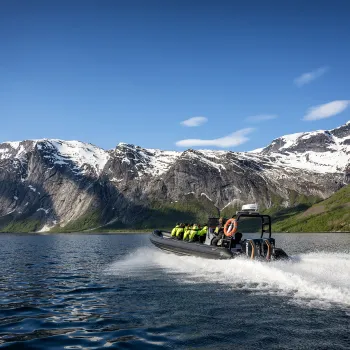En ribbåt med passasjerer i redningsvester suser over fjorden med snødekte fjell og blå himmel i bakgrunnen.