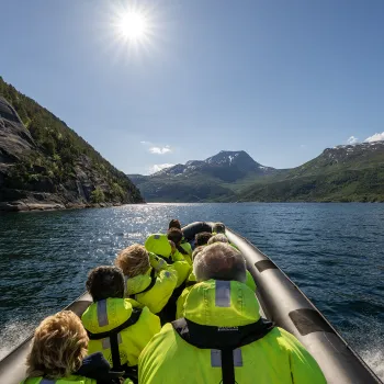Passasjerer i gule flytedrakter sitter i en RIB-båt på blå fjord, med fjell og sol på himmelen.