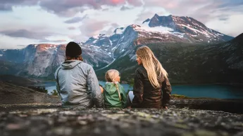 Tre personer sitter på en steinrygg og ser mot et fjellmassiv med snøflekker og en innsjø under rosa skyer.