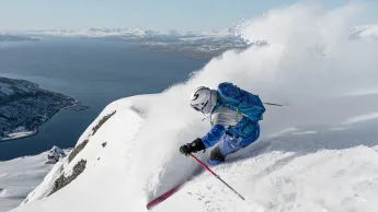 En skikjører i Narvikfjellet med hjelm og blå sekk svinger i puddersnø på fjellside, med fjord og snødekte fjell i bakgrunnen.