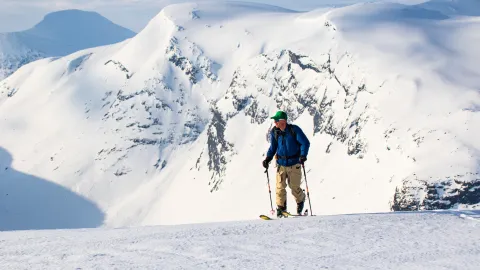 En skiløper med ryggsekk og staver står på en snødekt rygg, med bratte fjell og blå himmel i bakgrunnen.