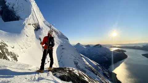 En skikjører med ryggsekk og ski på ryggen står på en snødekt fjelltopp med sol over fjord og fjell.
