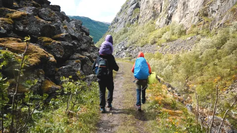 To turgåere med barn på rygg, med ryggsekker går på en smal sti i en fjelldal med steinur, trær og bratte fjellsider.