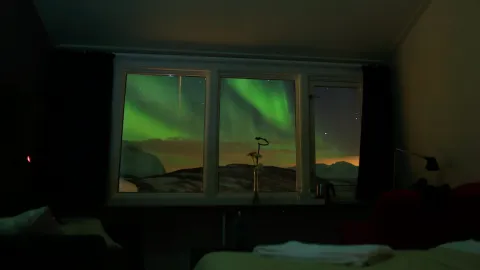 Grønt nordlys og stjernehimmel sett gjennom tre vindusruter, med mørkt rominteriør og seng i forgrunnen.